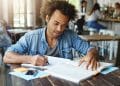 Homem de camisa jeans escreve em caderno enquanto consulta um livro aberto sobre mesa de madeira em ambiente de cafeteria