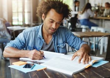 Homem de camisa jeans escreve em caderno enquanto consulta um livro aberto sobre mesa de madeira em ambiente de cafeteria
