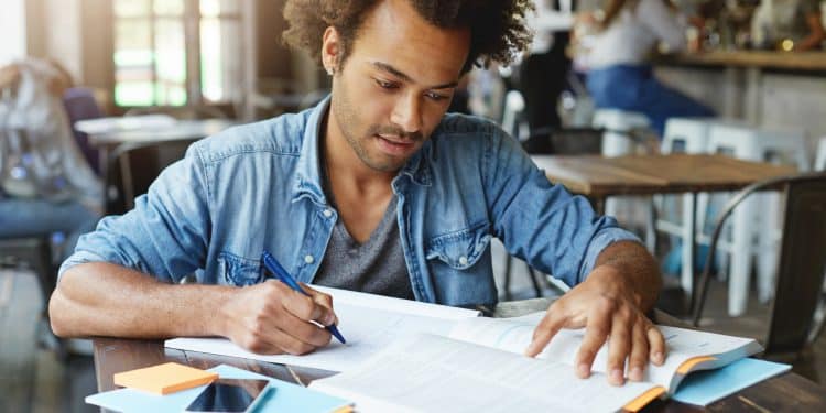Homem de camisa jeans escreve em caderno enquanto consulta um livro aberto sobre mesa de madeira em ambiente de cafeteria