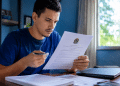 Homem de camiseta azul lê documento oficial com Brasão da República, segurando caneta, em home office com laptop.