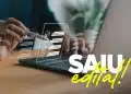 Mão segurando caneta stylus sobre laptop com ícones holográficos de checklist e texto "SAIU edital!" em destaque.