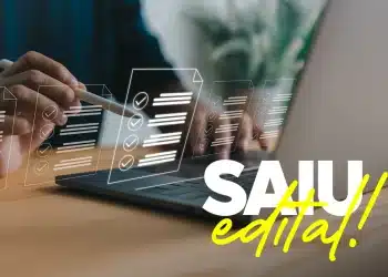 Mão segurando caneta stylus sobre laptop com ícones holográficos de checklist e texto "SAIU edital!" em destaque.