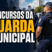 Agentes da Guarda Municipal em formação, usando uniformes azuis com coletes táticos e quepes, em área urbana.