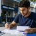 Jovem de camiseta azul marinho preenche formulários com caneta em frente ao prédio da Guarda Municipal.