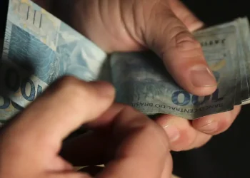 Mãos manuseando cédulas de cem reais brasileiros sobre fundo escuro em close-up