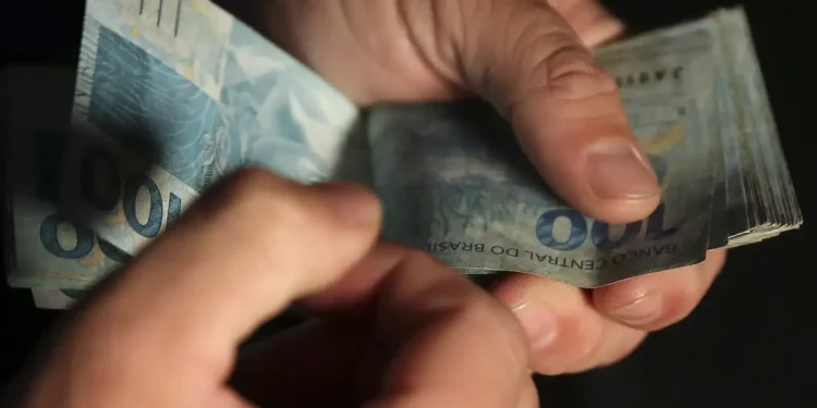 Mãos manuseando cédulas de cem reais brasileiros sobre fundo escuro em close-up