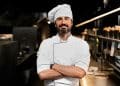 Chef de cozinha com uniforme branco e braços cruzados em cozinha