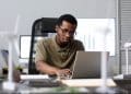 Homem de óculos trabalha concentrado em um notebook em um escritório, com maquetes de turbinas eólicas sobre a mesa