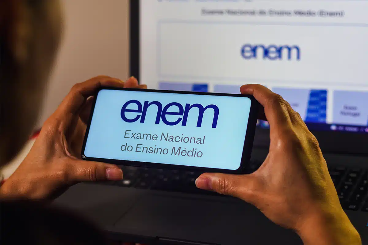 Mãos segurando smartphone horizontal exibindo logo do ENEM azul, com monitor mostrando Página do Participante ao fundo.