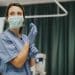 Enfermeira com scrub azul e máscara verde colocando luvas de látex azuis em quarto de hospital com cortina verde, simbolizando a enfermagem hospitalar.