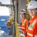 Duas profissionais sorrindo com uniforme laranja, capacetes brancos e óculos de segurança da Petrobras em plataforma offshore com o mar ao fundo