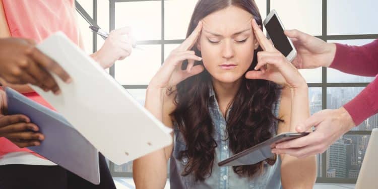 Mulher com olhos fechados pressionando as têmporas, cercada por mãos com celulares, papéis e tablets em ambiente de escritório