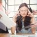 Mulher com olhos fechados pressionando as têmporas, cercada por mãos com celulares, papéis e tablets em ambiente de escritório