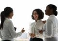 Três mulheres em traje social conversando e sorrindo em ambiente corporativo iluminado pela luz natural