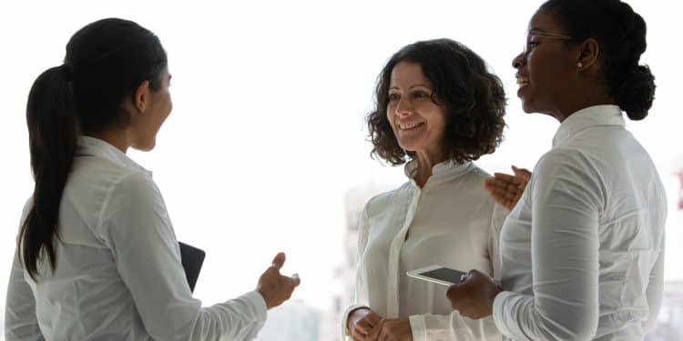 Três mulheres em traje social conversando e sorrindo em ambiente corporativo iluminado pela luz natural