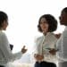 Três mulheres em traje social conversando e sorrindo em ambiente corporativo iluminado pela luz natural