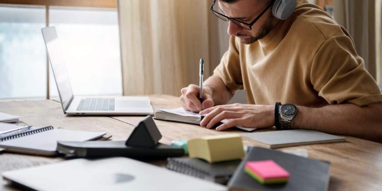 Estudante de fone de ouvido escreve em caderno ao lado de notebook em uma mesa de estudos, representação da rotina de preparação para o Enem com apoio de ferramentas de inteligência artificial.