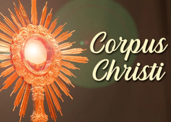 Ostensório dourado com raios solares exibindo hóstia consagrada e texto "Corpus Christi" em fundo marrom