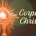 Ostensório dourado com raios solares exibindo hóstia consagrada e texto "Corpus Christi" em fundo marrom