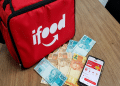 Bolsa térmica vermelha do iFood ao lado de notas de real e um celular com o aplicativo aberto sobre uma mesa