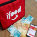 Bolsa térmica vermelha do iFood ao lado de notas de real e um celular com o aplicativo aberto sobre uma mesa