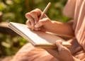 Mãos femininas segurando caderno espiral e escrevendo com caneta em ambiente externo iluminado pelo sol
