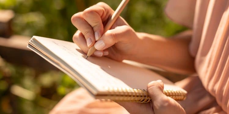 Mãos femininas segurando caderno espiral e escrevendo com caneta em ambiente externo iluminado pelo sol