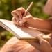 Mãos femininas segurando caderno espiral e escrevendo com caneta em ambiente externo iluminado pelo sol