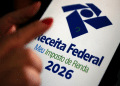 Mão com unhas vermelhas apontando para tela do celular exibindo logo da Receita Federal e Meu Imposto de Renda 2026.