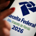 Mão com unhas vermelhas apontando para tela do celular exibindo logo da Receita Federal e Meu Imposto de Renda 2026.