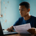 Jovem usa um notebook e segura um documento à mesa e, ao redor, um cômodo residencial de paredes azuis