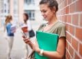 Estudante encostada em parede de tijolos consulta o celular enquanto segura caderno verde em pátio de instituição de ensino