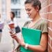 Estudante encostada em parede de tijolos consulta o celular enquanto segura caderno verde em pátio de instituição de ensino