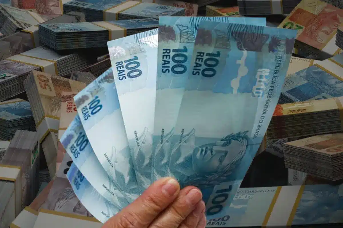Mão segurando notas de R$ 100, com maços de dinheiro ao fundo, representando o pagamento do Abono Salarial PIS/Pasep para trabalhadores formais.