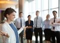 Executiva de blazer branco gesticula enquanto fala para cinco colaboradores sorridentes em lobby empresarial iluminado, simbolizando o curso Líder e Gestor de Equipes do Pensar Cursos.