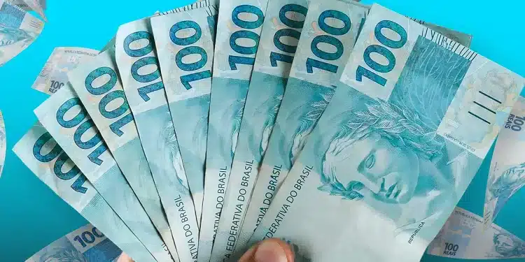 Mão segurando leque de notas de R$ 100 reais sobre fundo azul, representando a média salarial recorde do trabalhador brasileiro