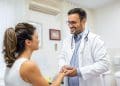 Médico de jaleco branco e estetoscópio sorri e segura as mãos de paciente em consultório médico iluminado, simbolizando o título de doutor dado à profissão.