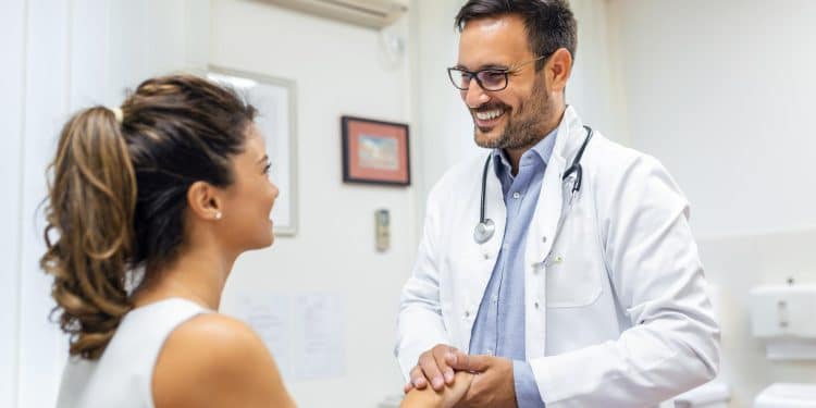 Médico de jaleco branco e estetoscópio sorri e segura as mãos de paciente em consultório médico iluminado, simbolizando o título de doutor dado à profissão.