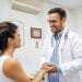 Médico de jaleco branco e estetoscópio sorri e segura as mãos de paciente em consultório médico iluminado, simbolizando o título de doutor dado à profissão.