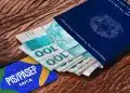 Carteira de Trabalho azul com cédulas de cem reais ao lado de cartão do PIS Pasep da Caixa sobre mesa de madeira