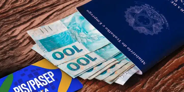 Carteira de Trabalho azul com cédulas de cem reais ao lado de cartão do PIS Pasep da Caixa sobre mesa de madeira