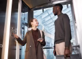 Duas pessoas conversando dentro de um elevador de vidro em prédio moderno