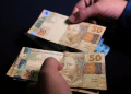 Mãos segurando cédulas de cinquenta reais com fundo escuro destacando o valor das notas brasileiras