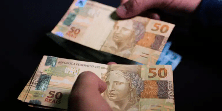 Mãos segurando cédulas de cinquenta reais com fundo escuro destacando o valor das notas brasileiras