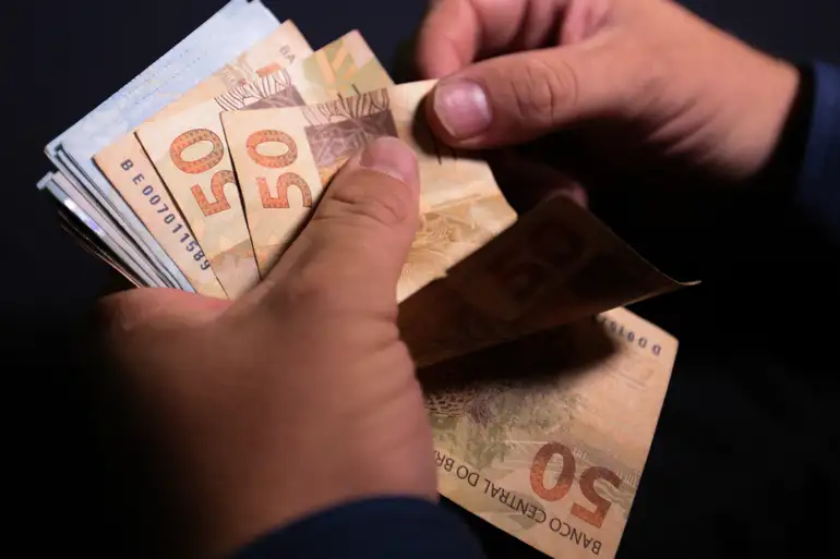 Mãos masculinas contando cédulas de cinquenta reais sobre fundo escuro