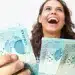 Mulher sorrindo segura notas de R$ 100 ao representar pagamento de R$ 1.486 ou mais para brasileiros com CPF de finais de 0 a 9.