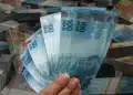 Alt text: Mão segurando notas de R$ 100 com maços de dinheiro ao fundo, representando pagamento de R$ 1.216 confirmado para CPF com final de 0 a 9