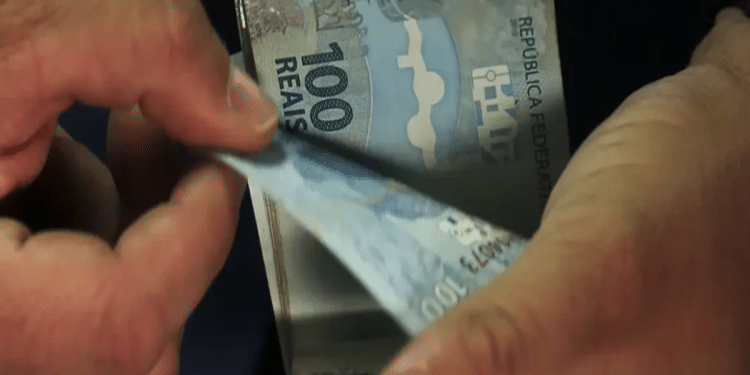 Mãos contando cédulas de R$ 100 reais brasileiros com fundo escuro, representando pagamento em dinheiro, simbolizando o pagamento do Pé-de-Meia.