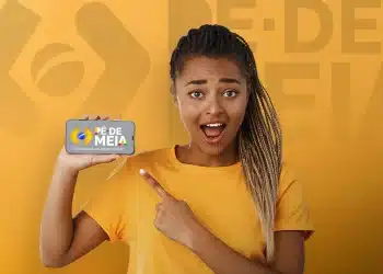 Estudante sorridente apontando para celular com a logo do Pé-de-Meia na tela em fundo amarelo, representando o pagamento do programa pelo Caixa Tem