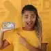 Estudante sorridente apontando para celular com a logo do Pé-de-Meia na tela em fundo amarelo, representando o pagamento do programa pelo Caixa Tem